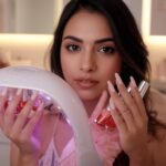 Beautiful woman with perfect pink and white ombre gel nails holding a modern LED nail lamp and glossy polish bottles on a bright bathroom vanity.