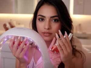 Beautiful woman with perfect pink and white ombre gel nails holding a modern LED nail lamp and glossy polish bottles on a bright bathroom vanity.